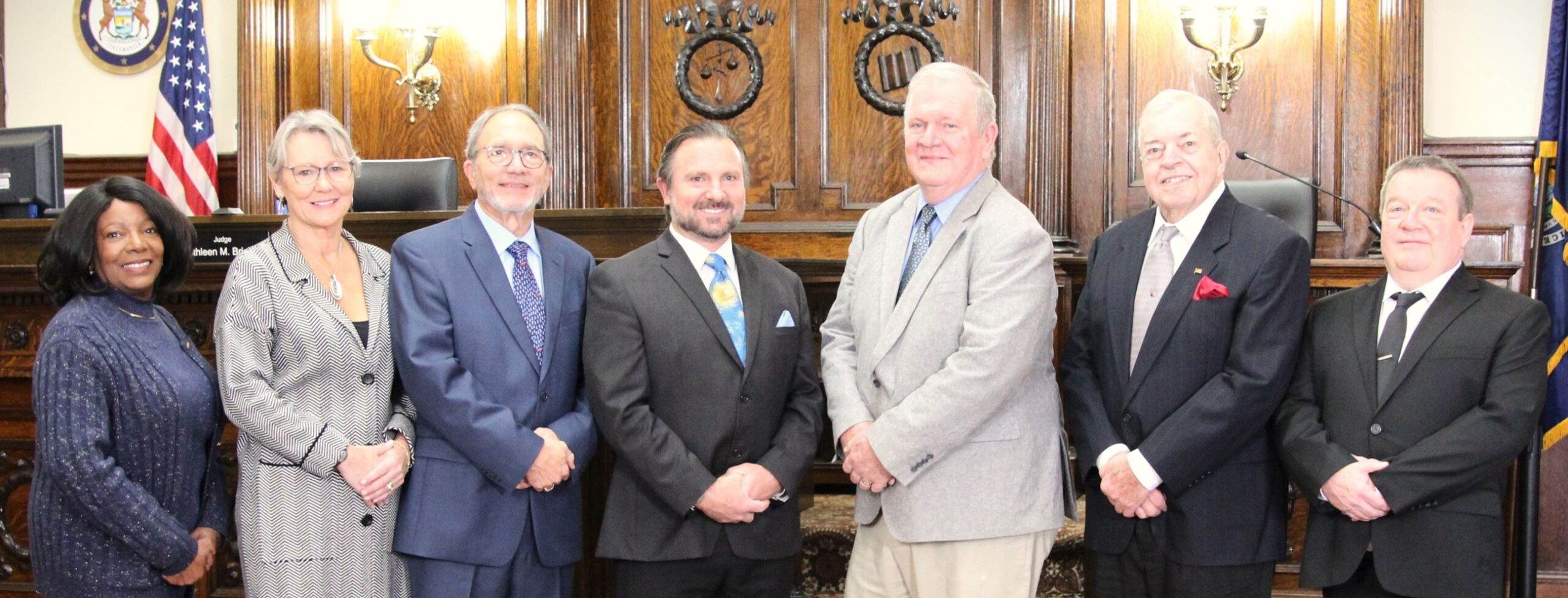 A group of six individuals stands together in a formal setting, likely a courtroom. They are dressed in professional attire, with some wearing suits and others in blazers. The background features wooden paneling and a judge's bench, along with an American flag. The individuals are smiling and appear to be posing for a photograph, suggesting a celebratory or official occasion.