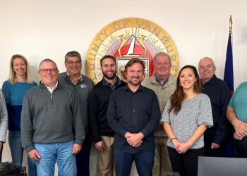 A group of eleven individuals is gathered in a room, standing in front of a large emblem that reads "County of Van Buren." The group consists of men and women of varying ages, dressed in casual to business attire. They are smiling and positioned closely together, with some holding pens and a bag in the foreground. Two flags are visible on either side of the emblem, and there is a camera mounted on the wall. The atmosphere appears friendly and collaborative.